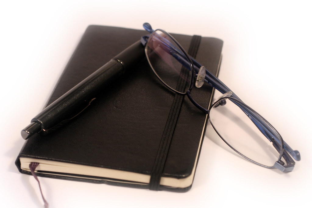 A closed notebook with a pen and reading glasses on a light surface